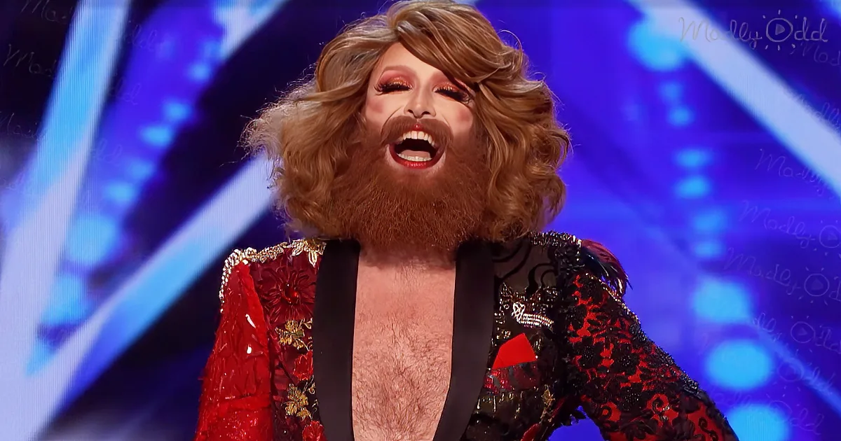 A person with a long, curly blonde wig and a full beard is wearing a red and black outfit with a gold pattern, standing...