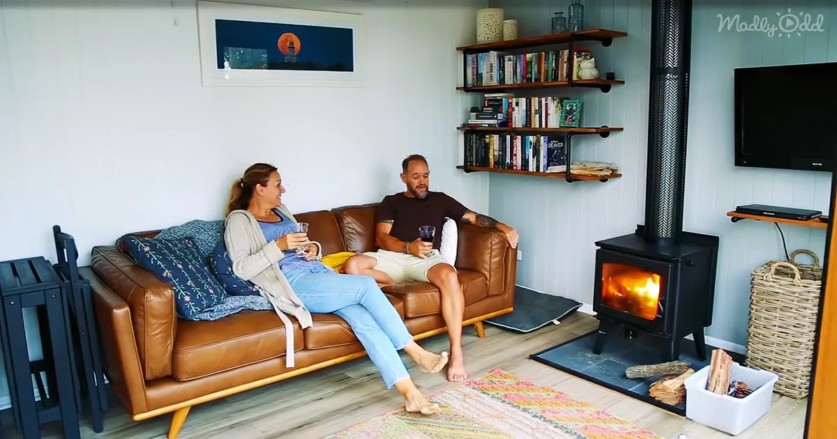 A couple sits on a brown leather couch in a cozy room with a wood-burning stove, books, and a TV.
