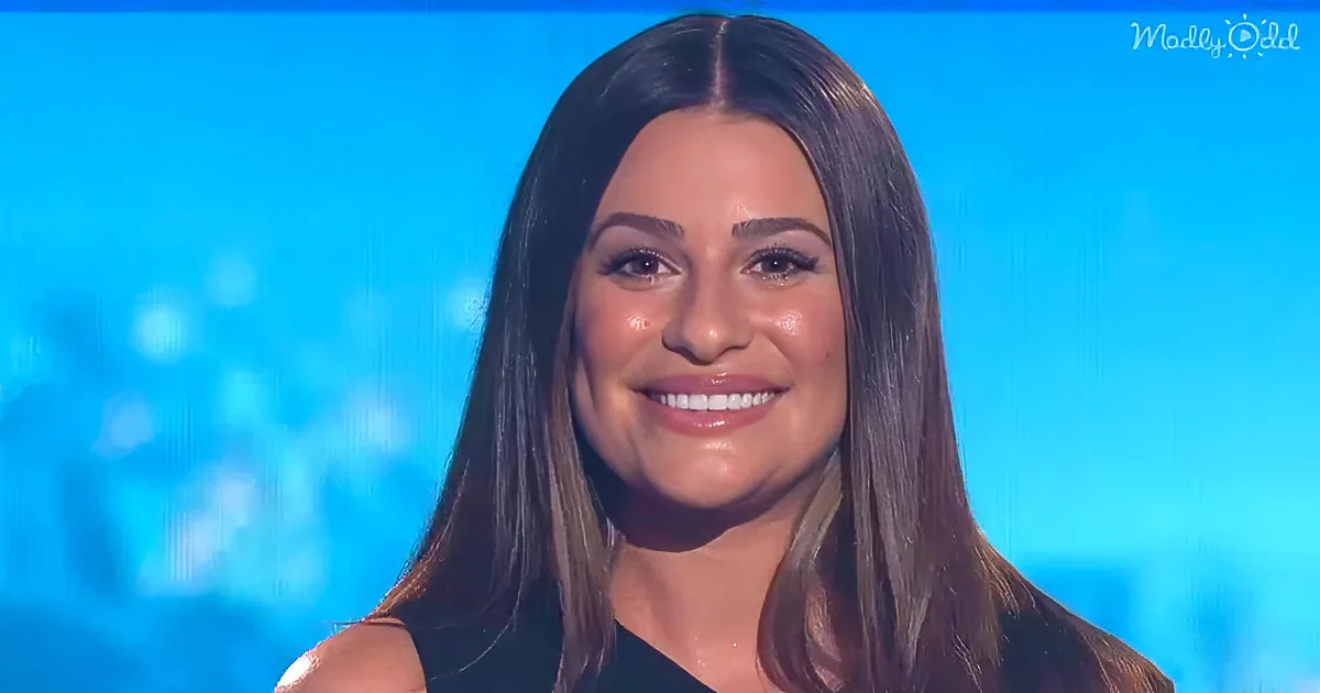A woman with long dark hair smiles, wearing a black top, against a blue background with blurred lights.