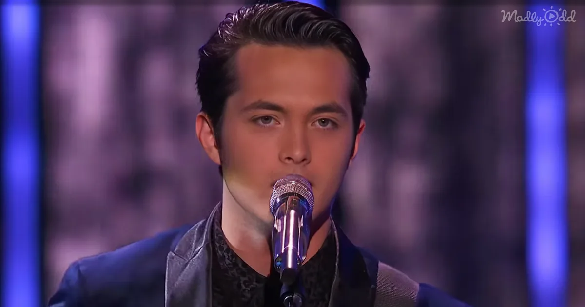 A man with dark hair and a microphone, wearing a dark jacket, is singing on stage with a purple background.