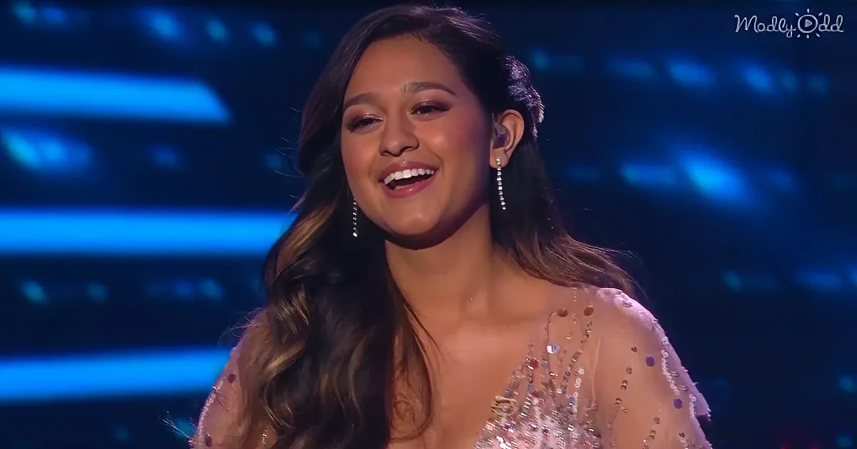 A woman with long, wavy hair and a beaded dress smiles on stage, wearing earrings.