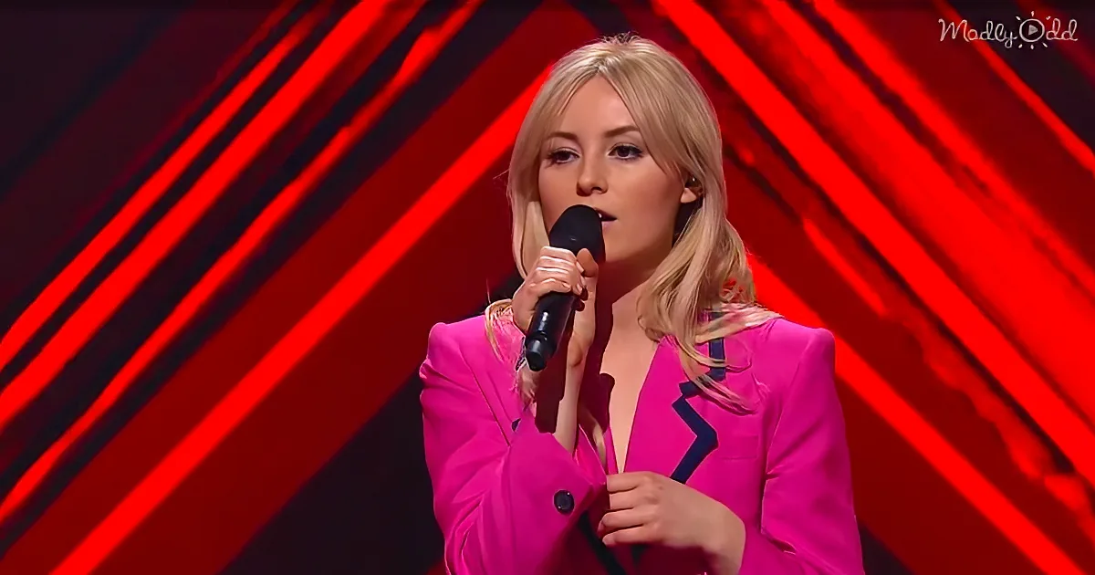 A woman in a bright pink suit sings into a microphone against a red, striped background.