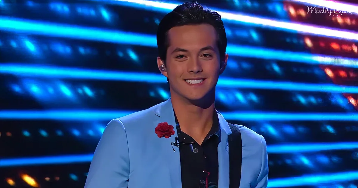 A man in a light blue suit with a red flower on his lapel smiles on a stage with blue lighting.