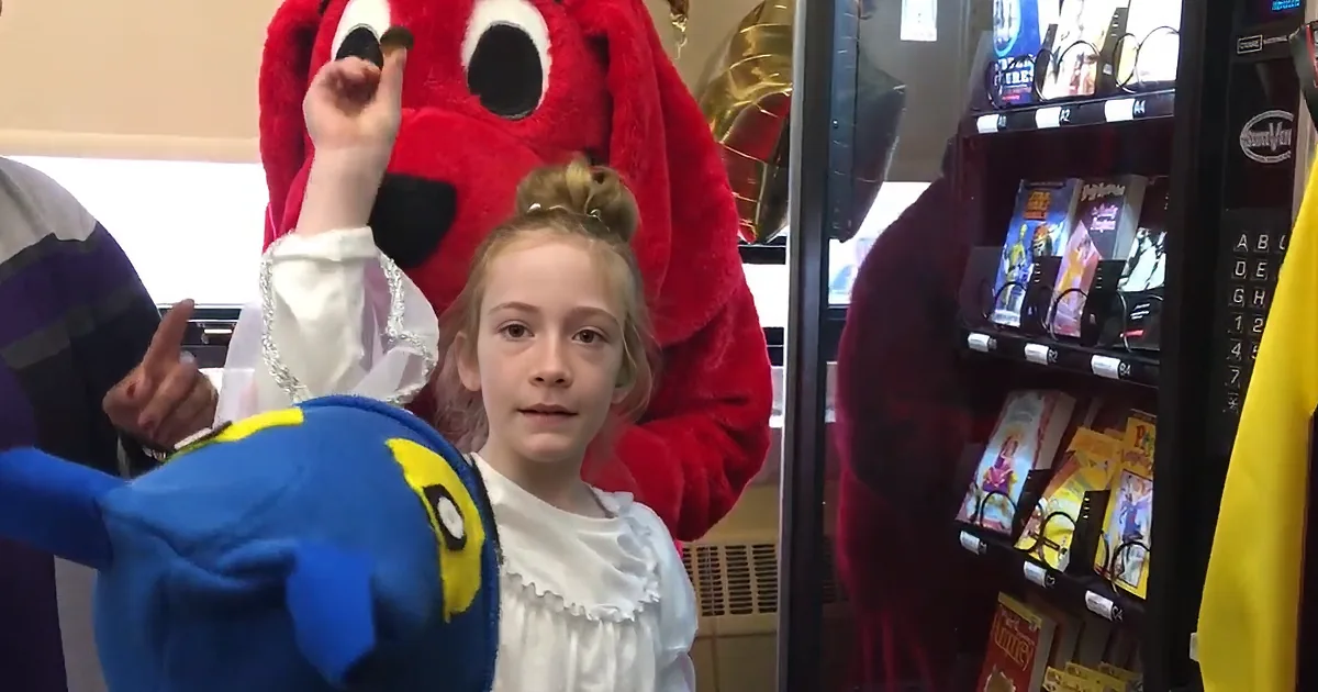 A young girl in a white dress stands in front of a red mascot, with a blue plush toy in the foreground.