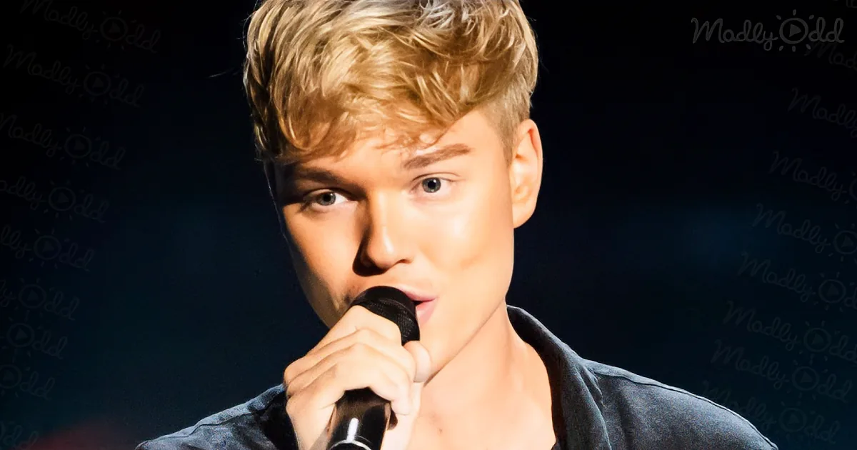 A person with blonde hair holding a microphone, wearing a dark shirt, against a dark background.