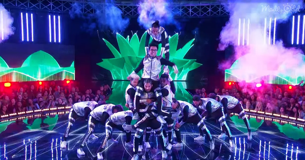 A group of performers in white and black outfits form a human pyramid on stage with vibrant lighting and smoke effects.