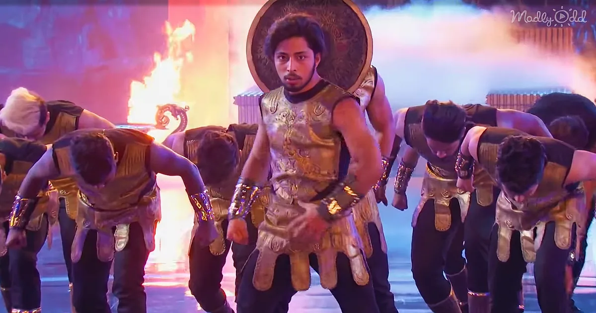 A man in golden armor stands confidently, surrounded by dancers in similar attire, with a fiery backdrop.