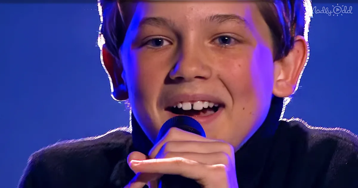 A young boy with short brown hair holds a microphone, singing with his eyes closed, under blue stage lighting.