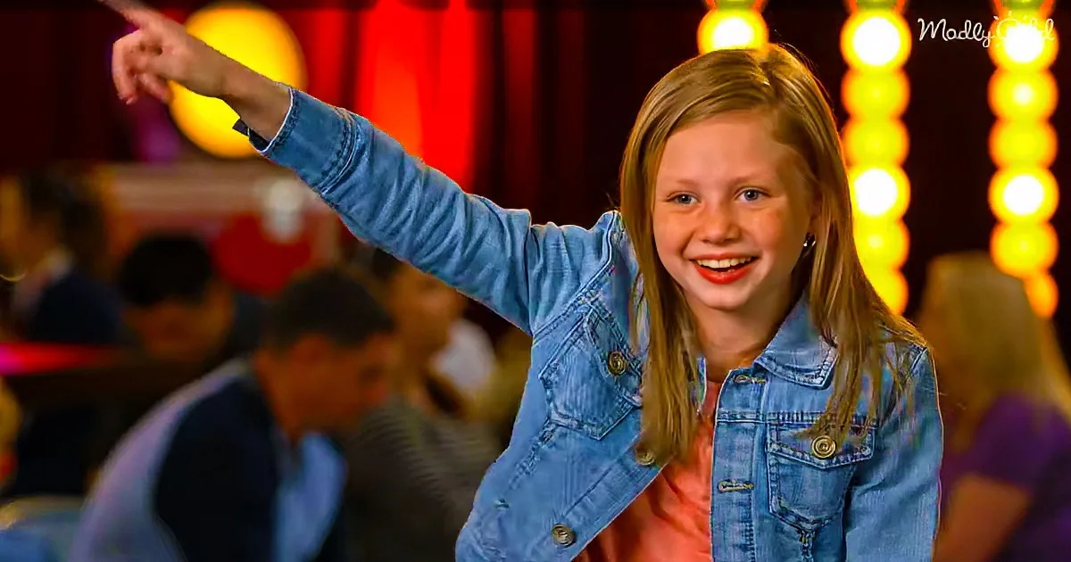 A girl in a denim jacket smiles and points to the left in a lively setting with bright lights.