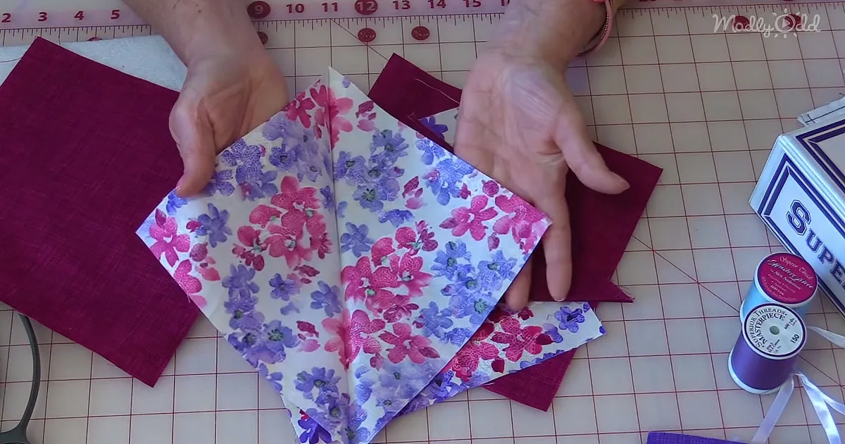 Hands fold floral fabric on a grid cutting mat with red and purple fabrics nearby.