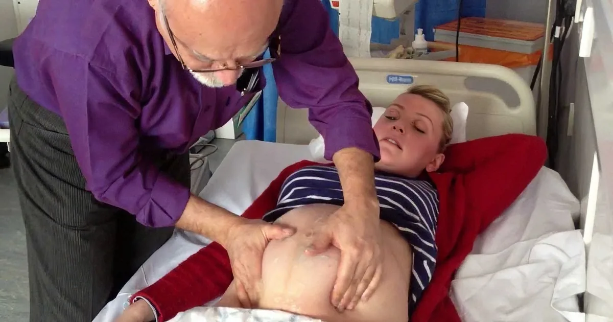 A man in a purple shirt is examining a person's abdomen in a hospital setting.