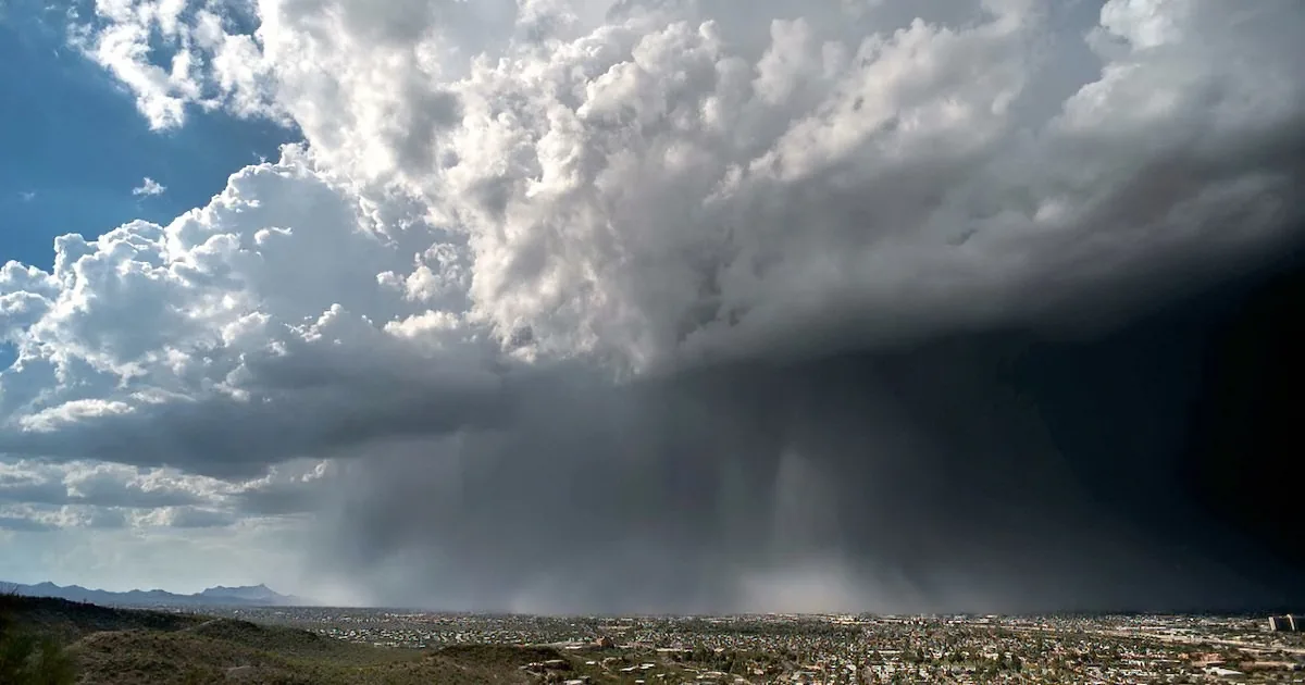 A massive dark storm cloud looms over a cityscape, with rain visibly falling from its base.