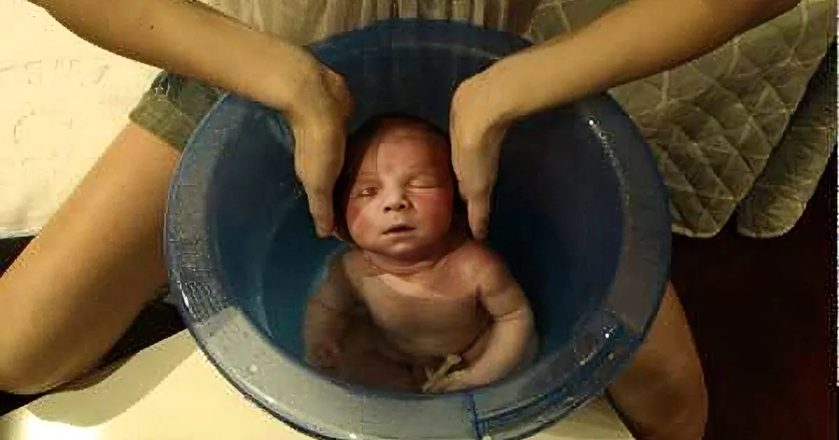 A newborn baby is being held in a blue bucket by two hands, appearing calm and content.