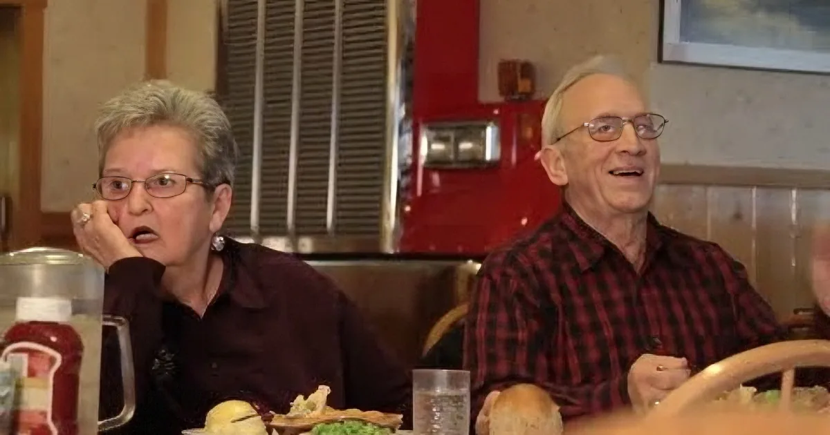 An elderly couple sits at a diner table, enjoying a meal together, with a red fire truck visible in the background.