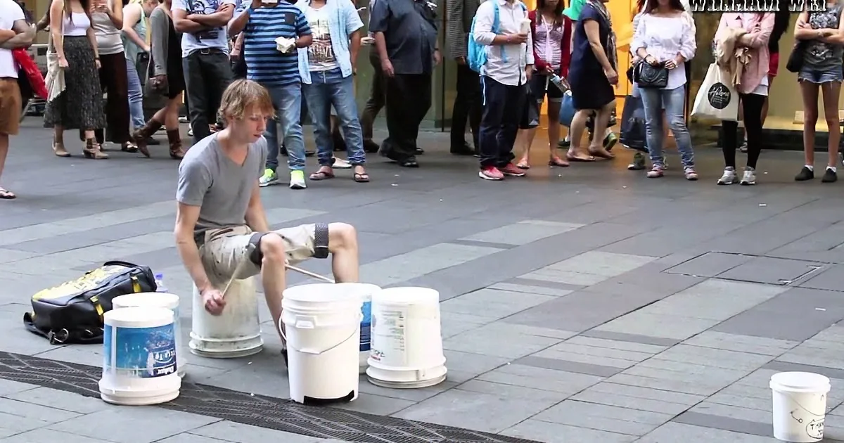 A street performer plays drums on buckets in a public square, surrounded by an audience.
