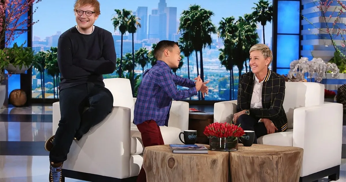 Ed Sheeran sits on a white chair, smiling, while a young boy and Ellen DeGeneres engage in conversation on a talk show set.