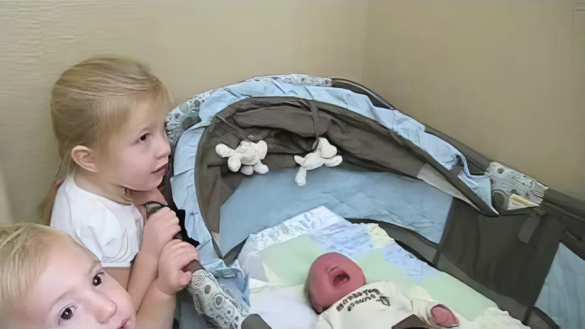 A young girl holds a microphone, looking at a newborn baby in a bassinet.