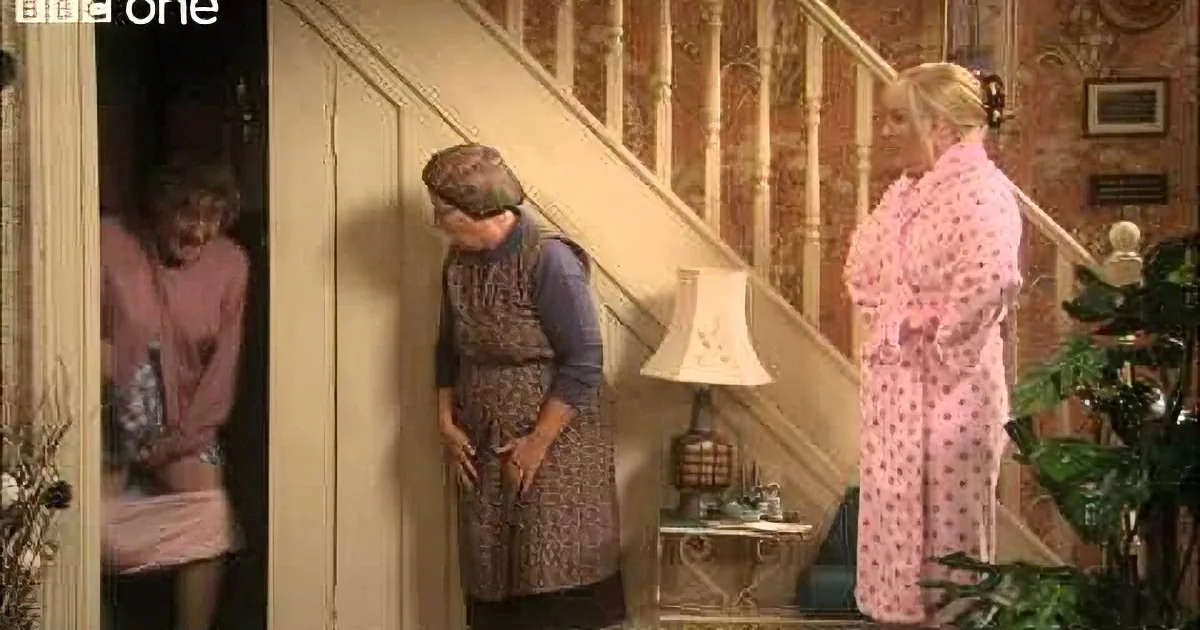 A woman in a pink robe stands near a staircase, while another woman in a patterned dress leans against the wall.