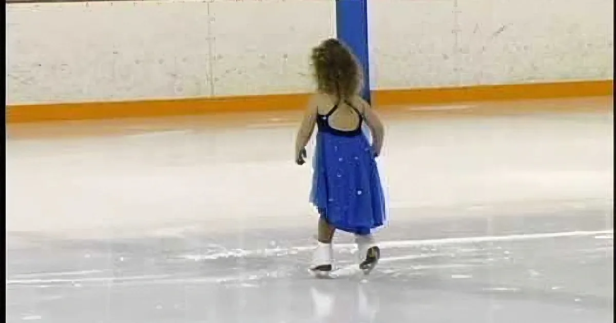 A child in a blue dress skates on an ice rink, facing away from the camera.