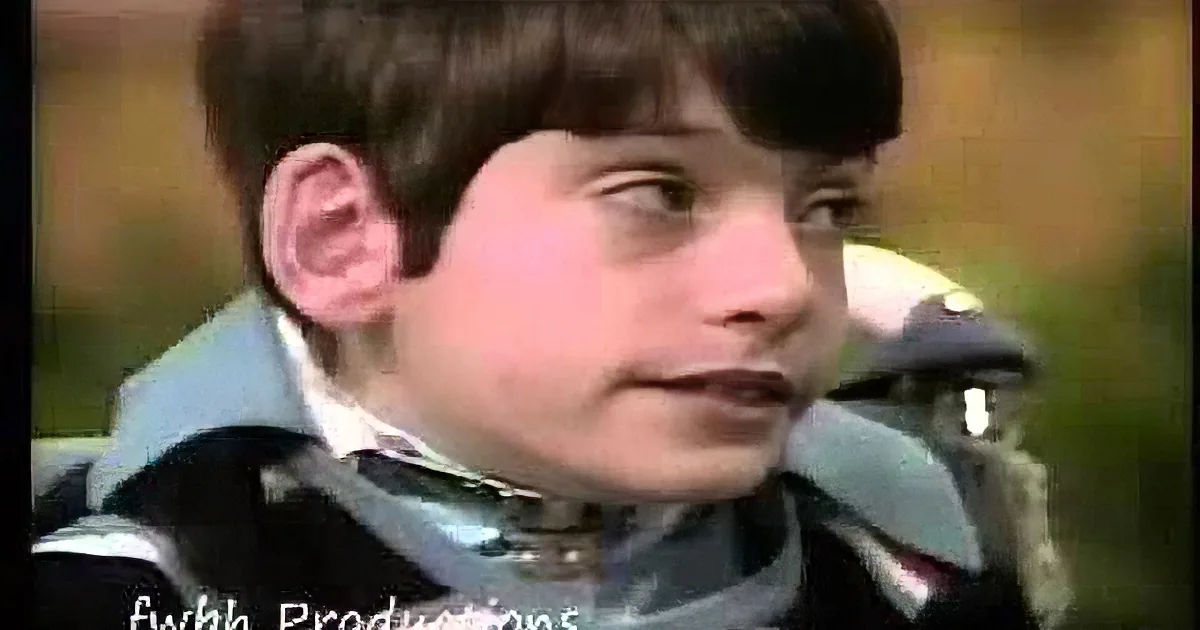 A young boy with short brown hair, wearing a striped shirt and a jacket, looks to the side with a serious expression.