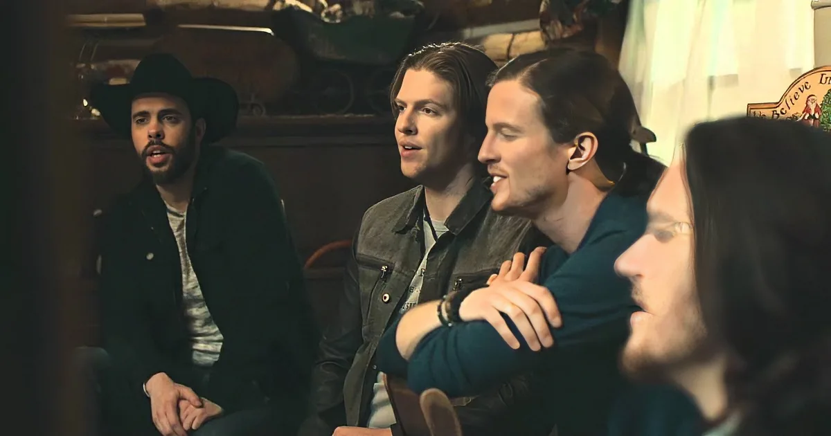 Three men in a rustic setting, one wearing a cowboy hat, engaged in conversation.