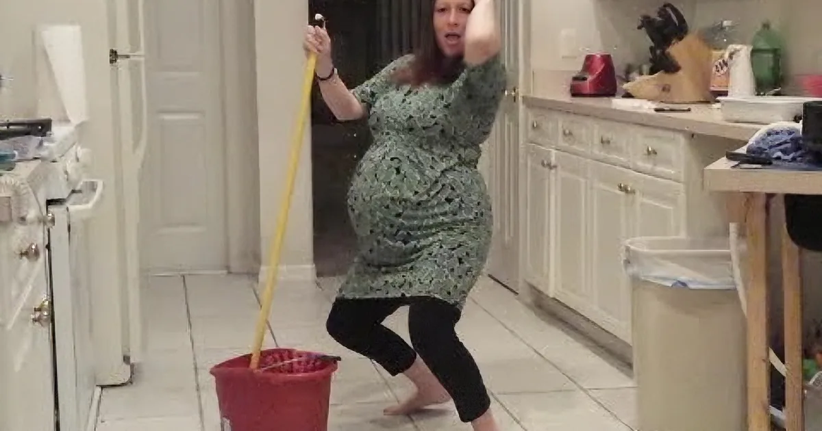A pregnant woman in a green dress is mopping the floor in a kitchen.