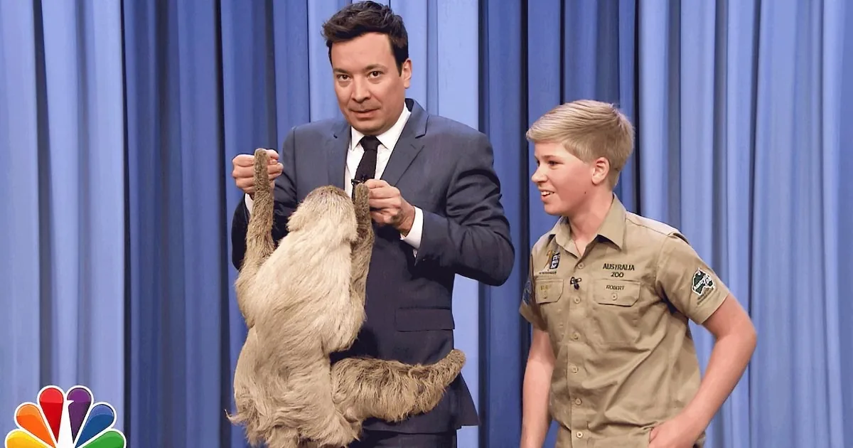 A man in a suit holds a sloth while another person in a zoo uniform looks on.