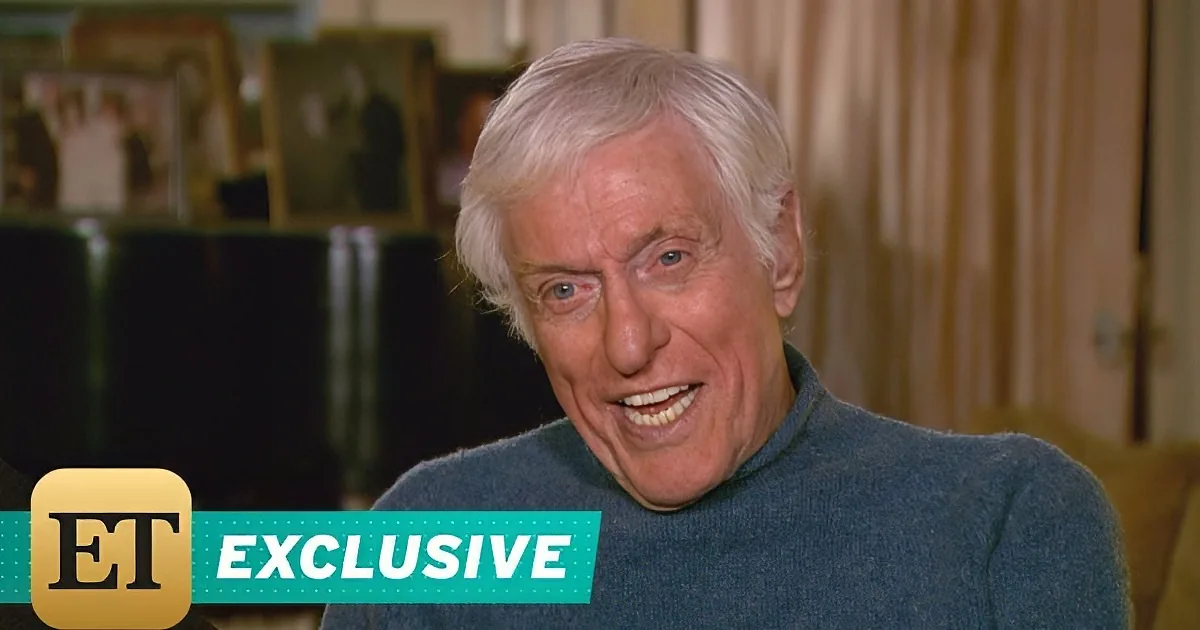 An older man with white hair smiles, wearing a blue turtleneck, in a softly lit room with framed pictures.