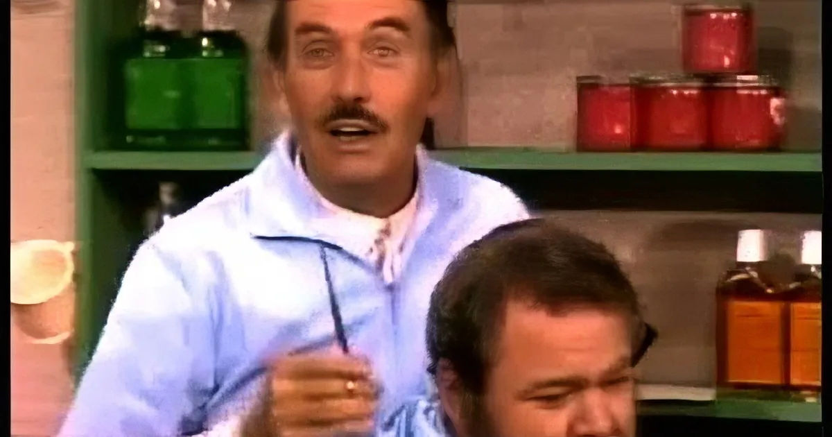A man in a white coat holds a pair of scissors, standing behind another man in a kitchen setting.