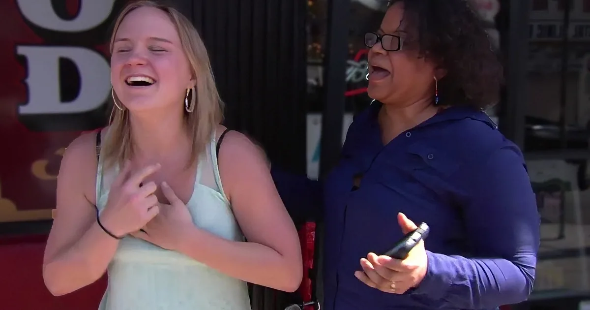 Two women are laughing outdoors, one in a white tank top, the other in a blue hoodie, holding a phone.