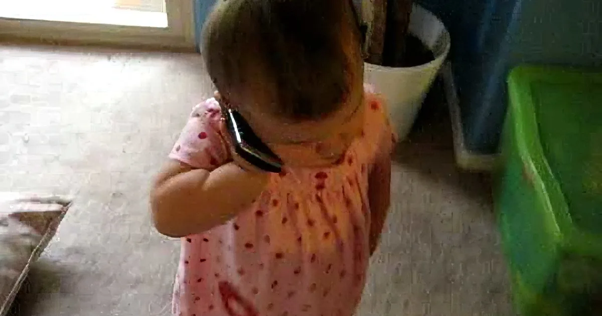 A toddler in a pink polka-dot dress holds a black object near their ear, possibly a phone.