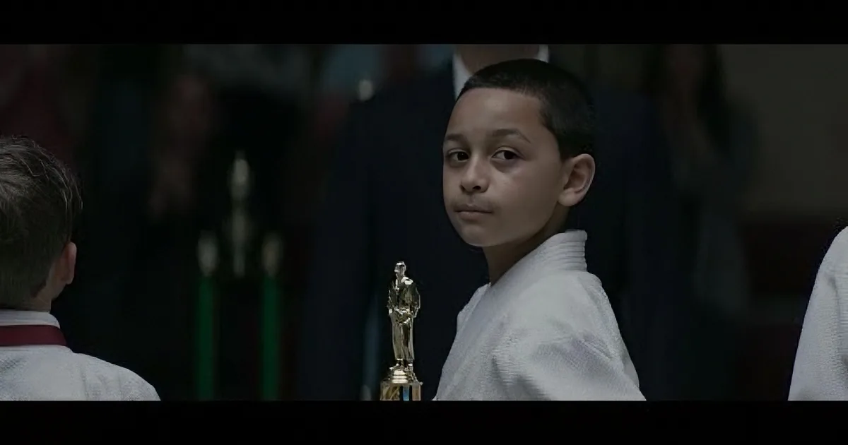 A young boy in a white karate uniform holds a trophy, looking to the side with a serious expression.
