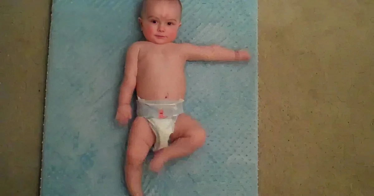 A baby in a diaper lies on a blue mat, arms outstretched, looking up.