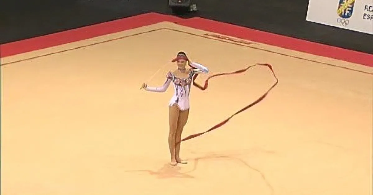A gymnast in a white leotard performs with a red ribbon on a beige floor.