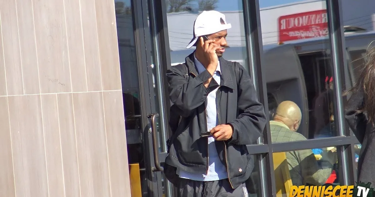 A man in a white cap and black jacket talks on his phone, exiting a building.
