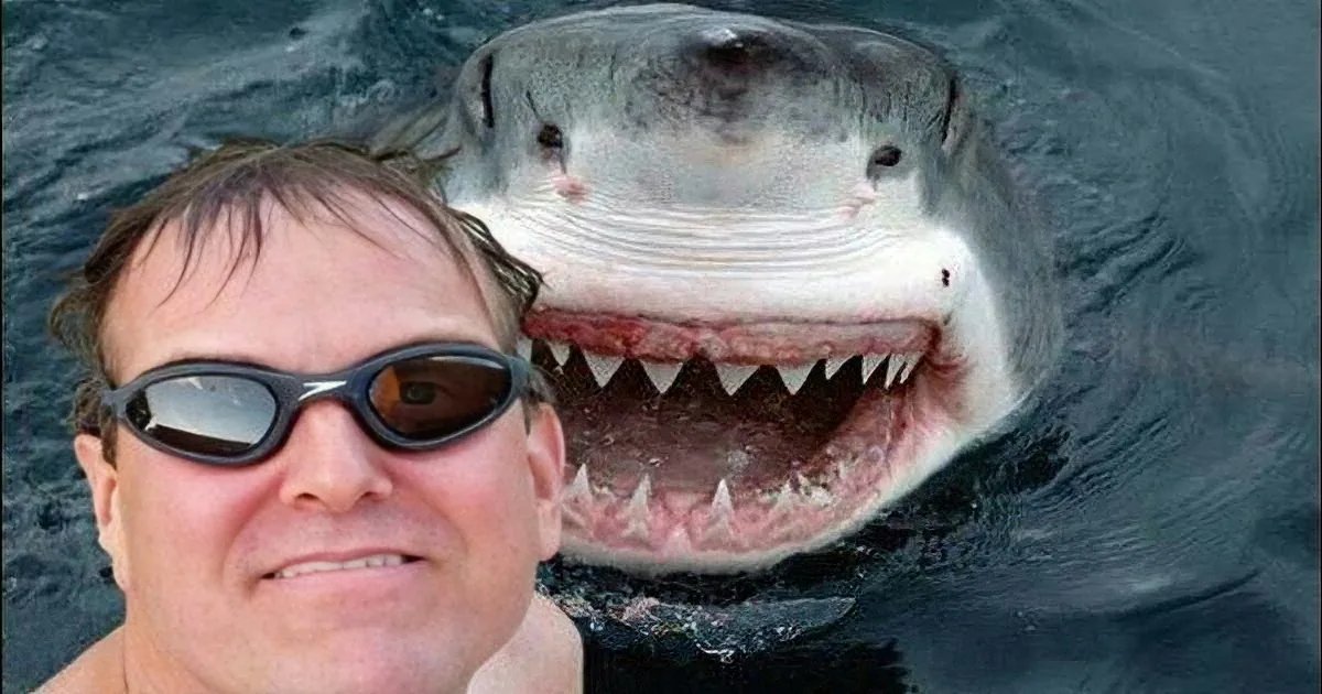 A man wearing goggles smiles next to a large shark with its mouth open.