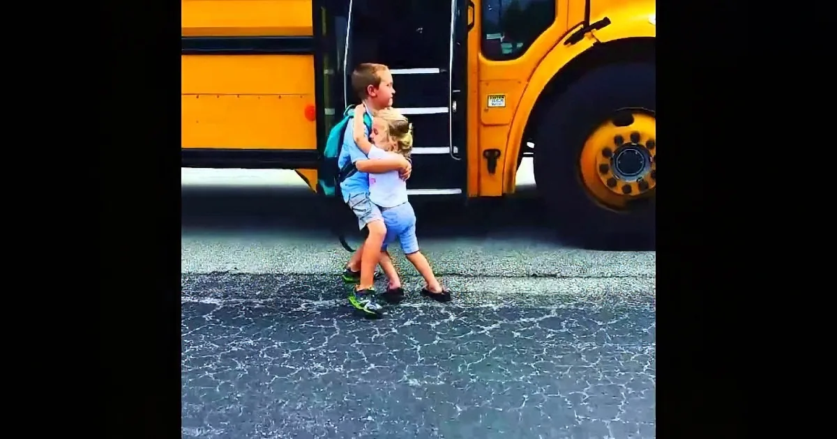 A boy and girl hug in front of a yellow school bus on a cracked asphalt surface.