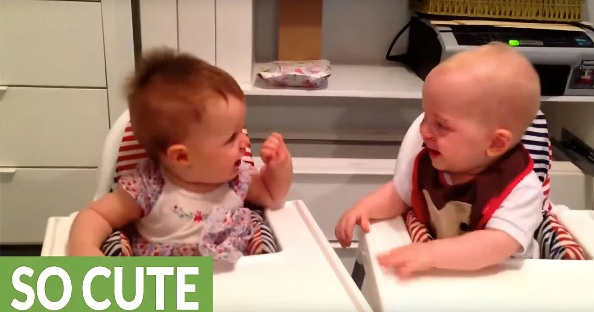 Two babies in high chairs, one in a floral bib, the other in a brown bib, interacting with each other.