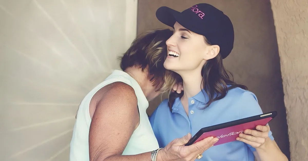 A woman in a blue shirt and cap hugs another woman while holding a pink tablet.