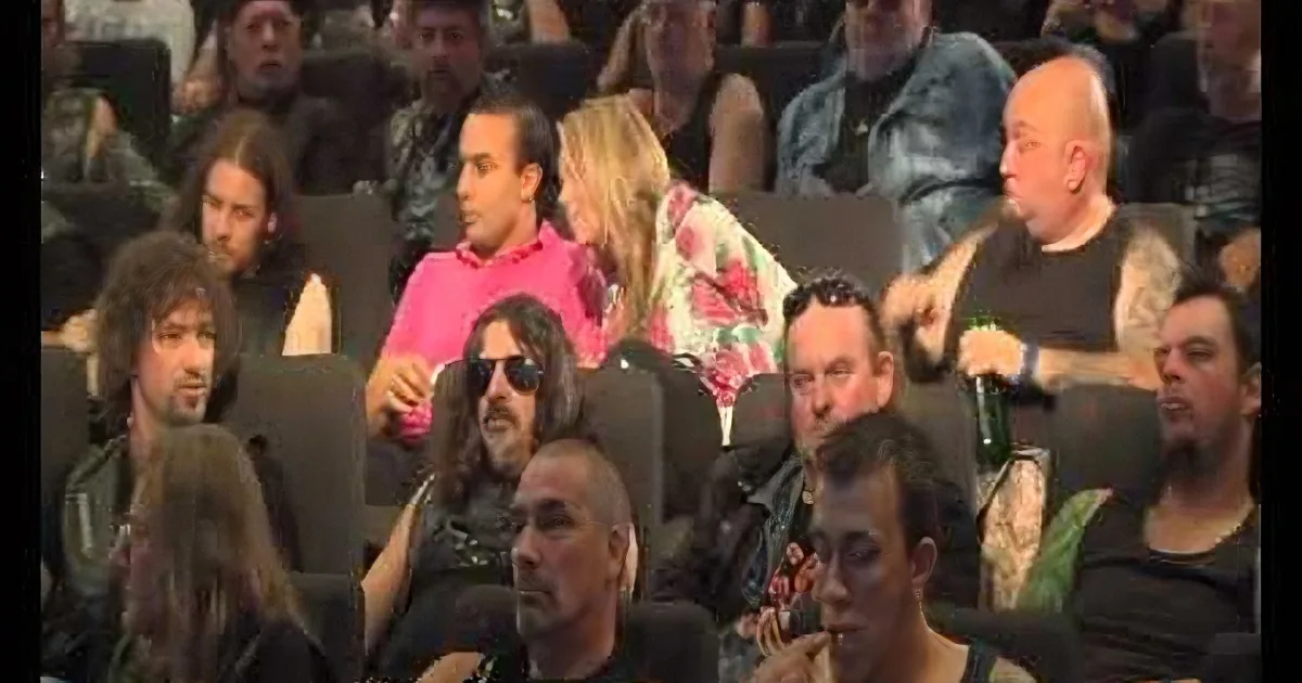 A man in a pink shirt sits in a crowded audience, surrounded by people in various attire, some holding drinks.