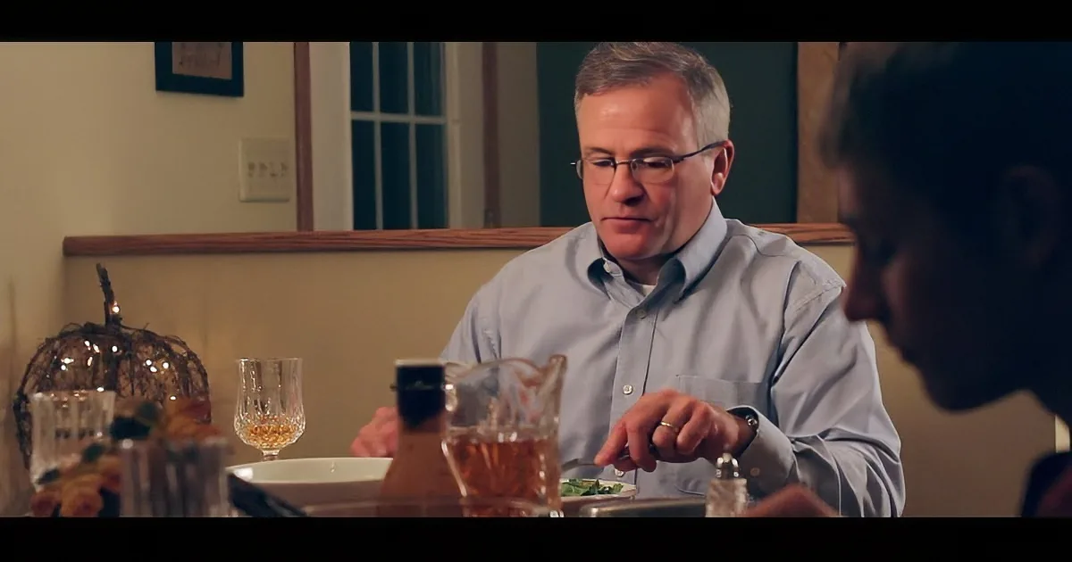 A man in a light blue shirt sits at a table, holding a fork, with a glass of wine and a pitcher nearby.