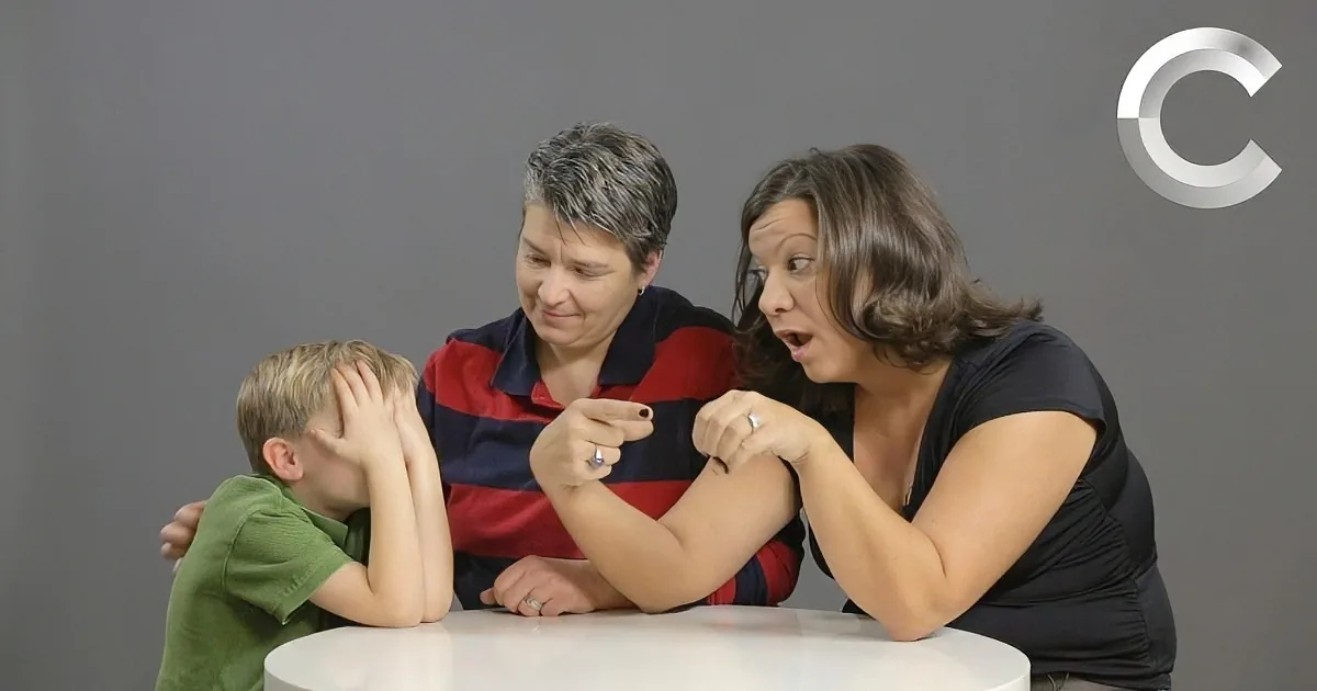 A woman in a red striped shirt is pointing at another woman in a black shirt while a child covers their face with their...