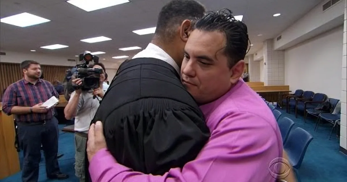 A man in a pink shirt hugs another in a black robe in a courtroom setting.