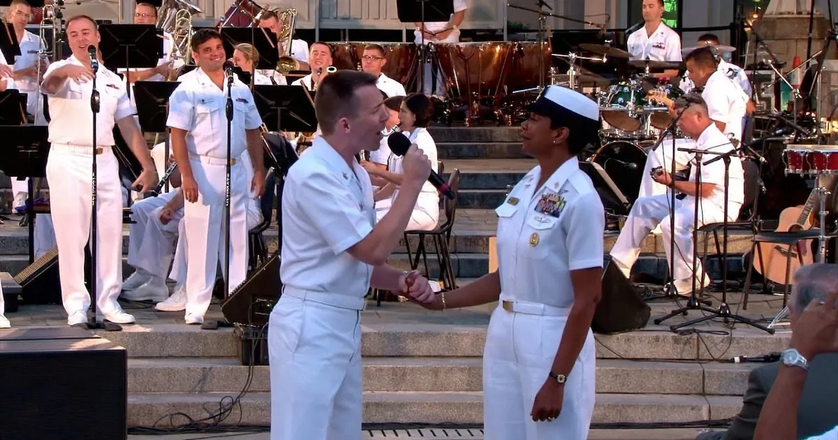 A man in white uniform shakes hands with a woman in uniform while a band plays in the background.