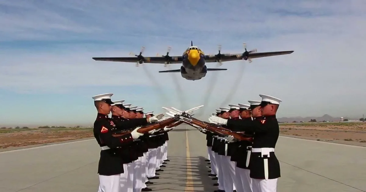 A military honor guard stands in formation, rifles raised, as a C-130 Hercules aircraft flies overhead.
