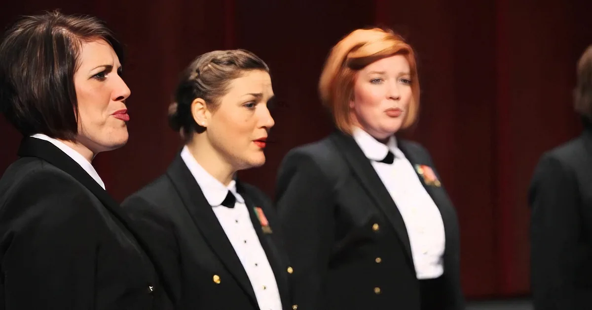Three women in formal black uniforms with white shirts and gold buttons stand in a row, facing left.
