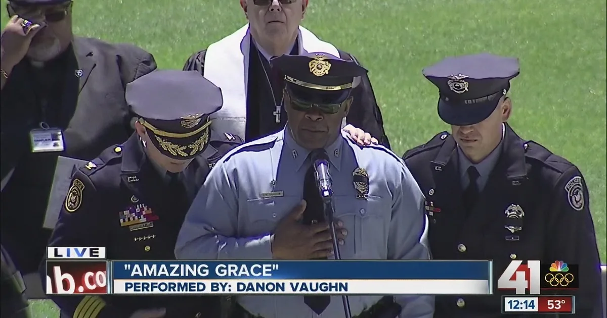 A police officer stands at a podium, singing "Amazing Grace," with other uniformed personnel behind him.