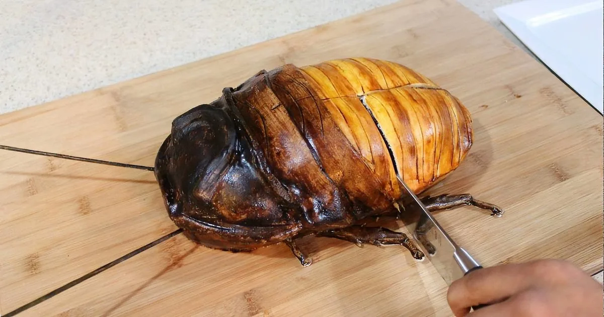 A roasted insect, possibly a cockroach, is being cut on a wooden cutting board with a knife.