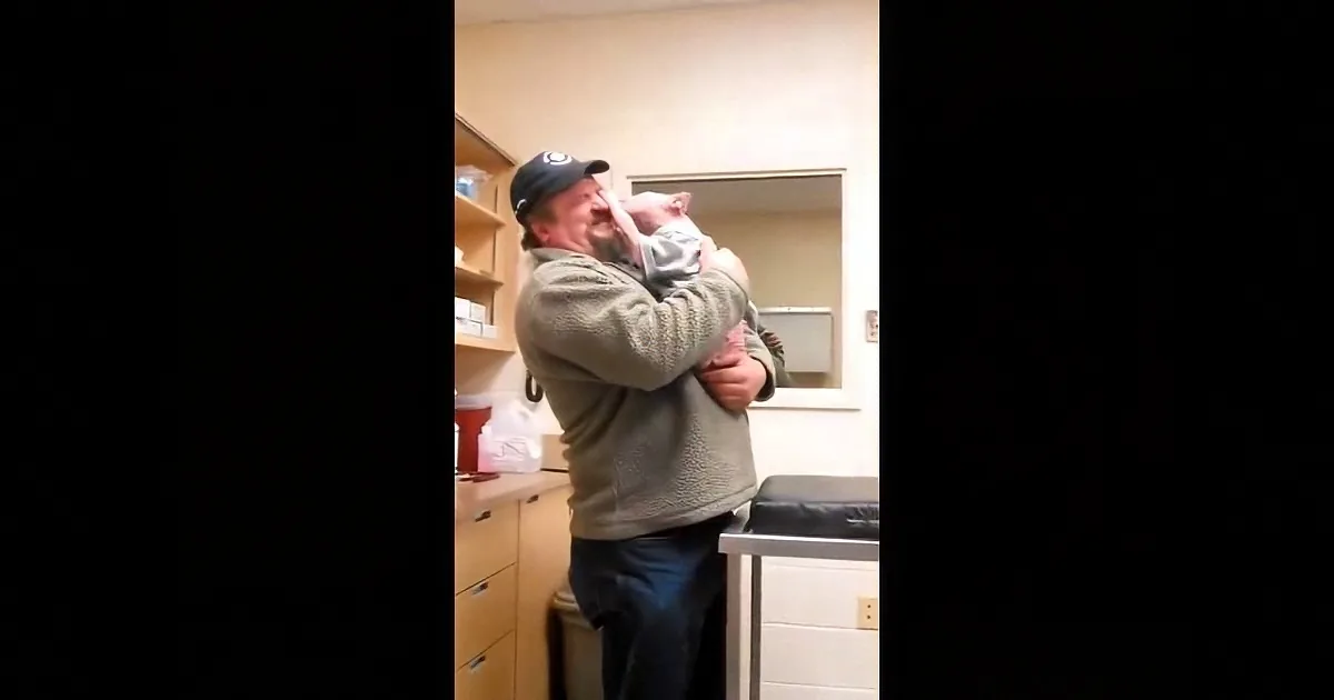 A man in a cap holds a baby, both smiling, in a room with shelves and a mirror.