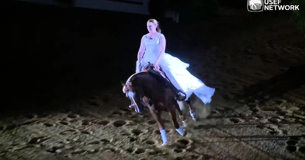 A woman in a white dress rides a horse in a dimly lit arena.